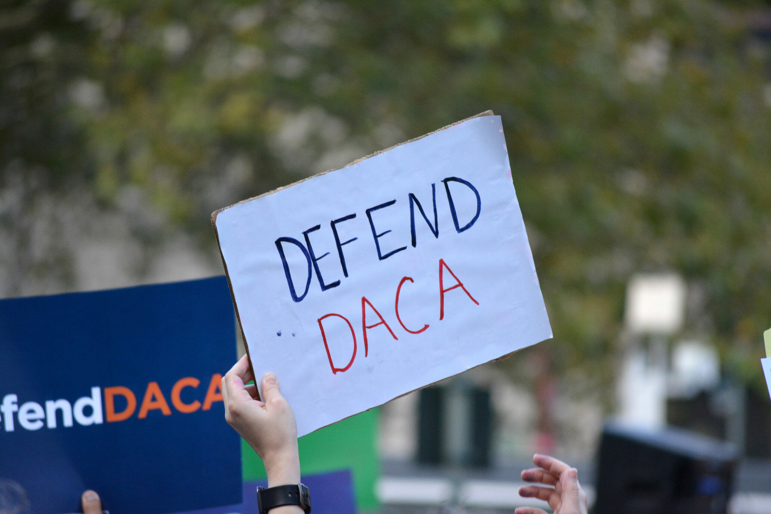 A hand holding a small white sign at a rally that says "Defend DACA"