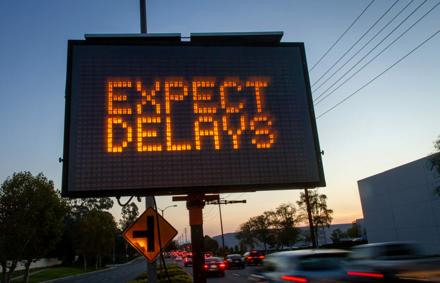 Lighted traffic sign reading "expect delays" in capital organe letters, with a dusk sky in the background and cars driving by in the foreground.